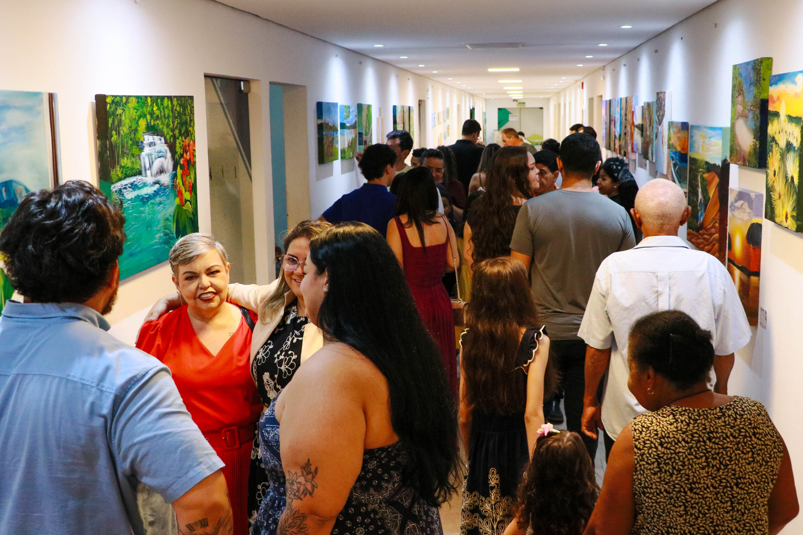 Visitantes conversam em salão com pinturas expostas nas paredes na mostra Janelas para o Mundo.
