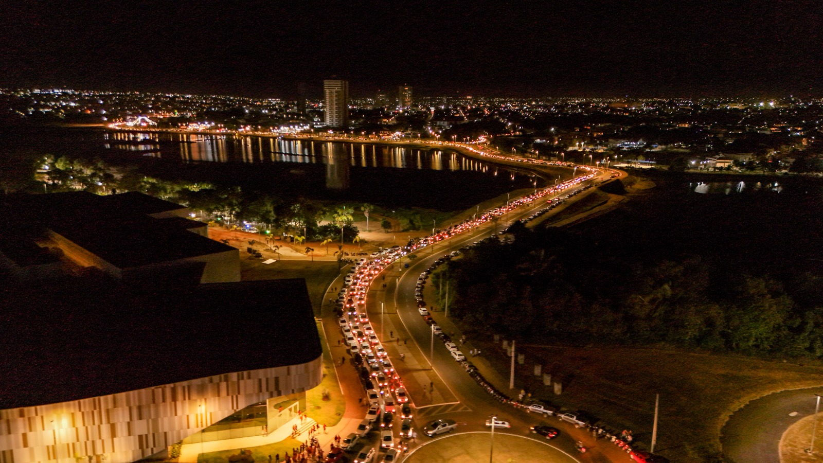 Vista aérea noturna de avenida à margem de lago em Araguaína, com fila de veículos.