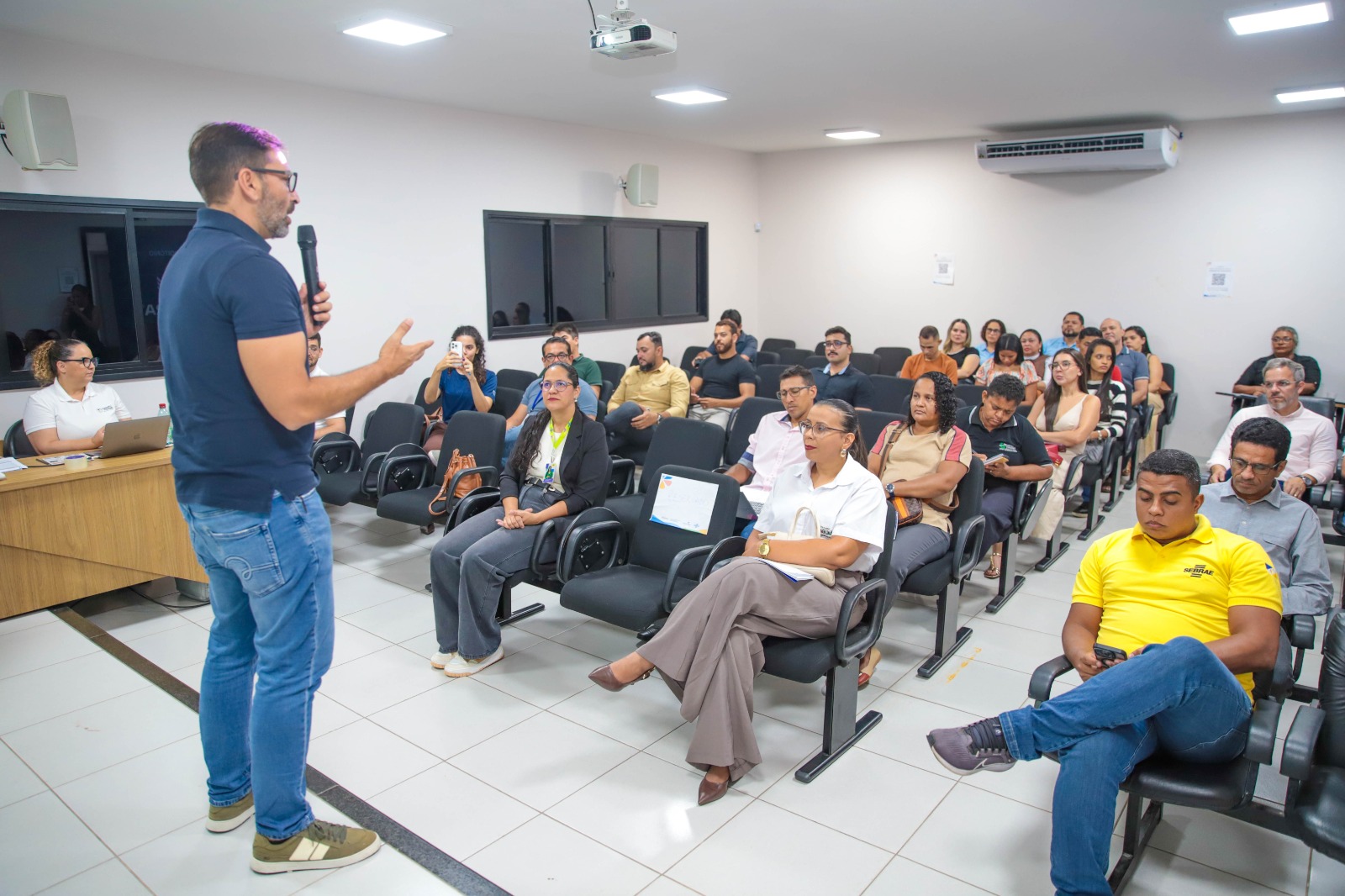Homem fala ao microfone diante de participantes sentados em auditório durante audiência sobre turismo.