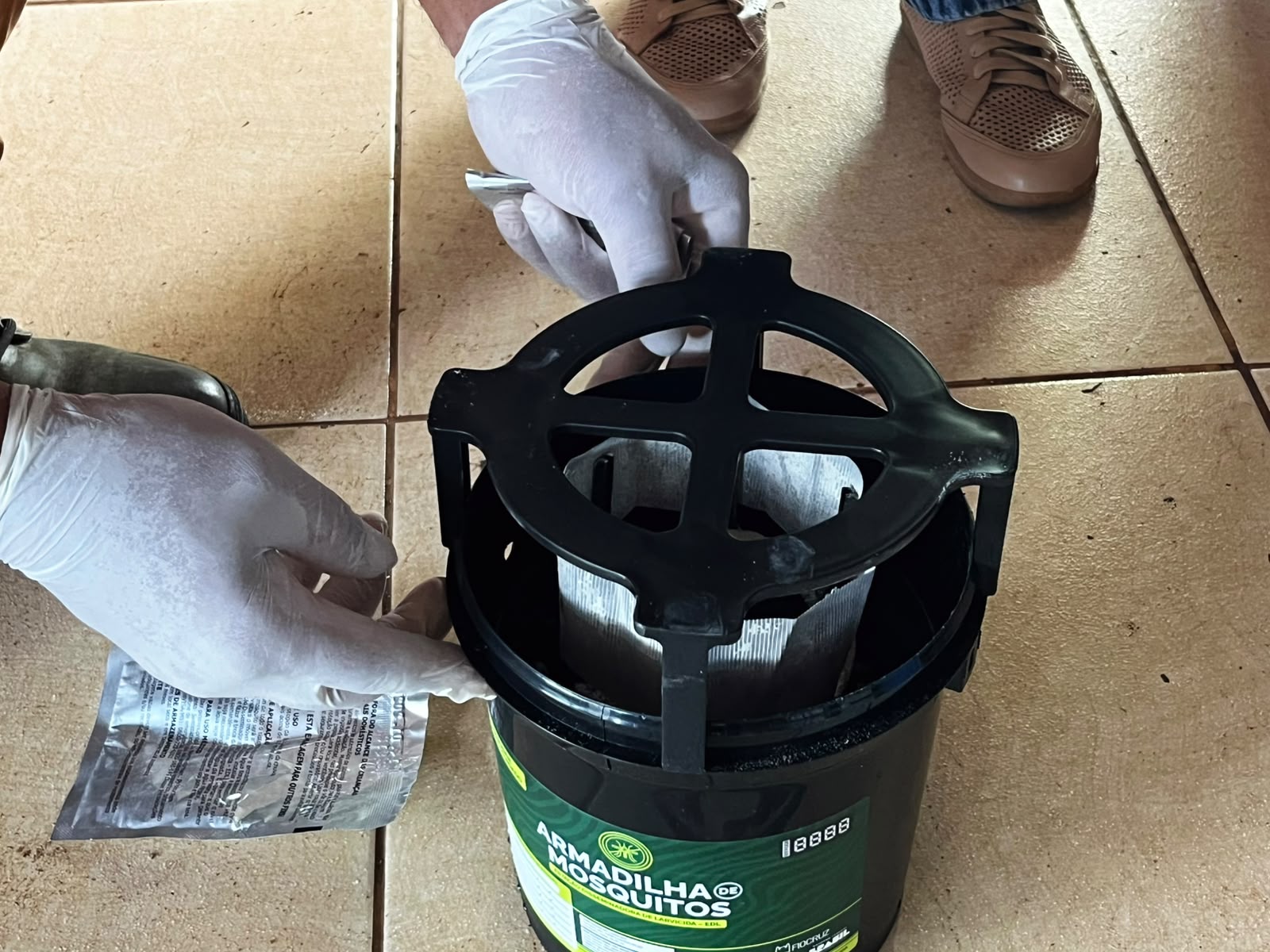 Mãos com luvas manipulam uma armadilha preta para mosquitos sobre piso cerâmico bege.