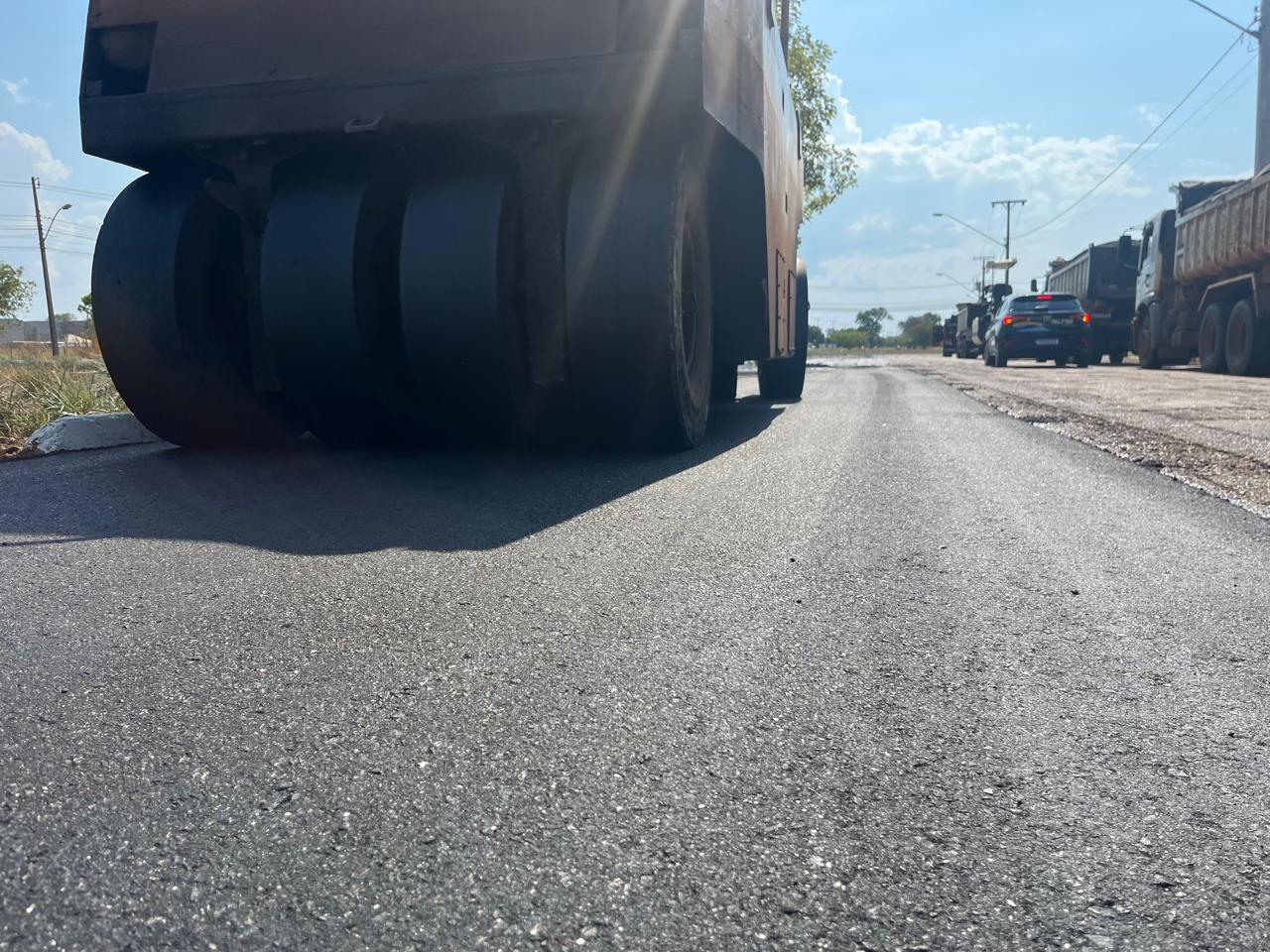 Rolo compactador sobre asfalto recém-pavimentado, com carro e caminhões ao fundo em uma via em obras.