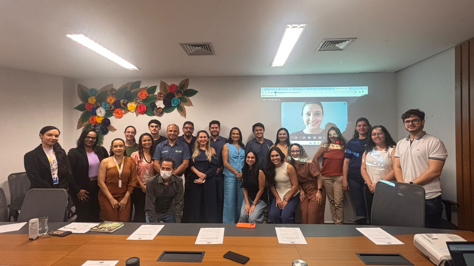 Participantes reunidos em sala com documentos sobre a mesa e videoconferência projetada na parede.