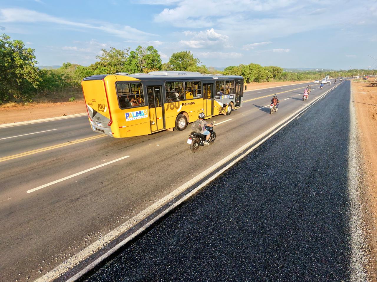Faixa recém-asfaltada ao lado de avenida em Palmas, separada por guia de concreto, com ônibus e motocicletas em circulação.